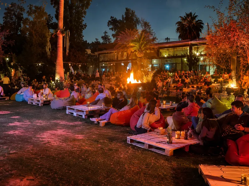 Bonfire cinema night: people on beanbags and pallet tables around a lit fire under the trees, waiting for the film to start.