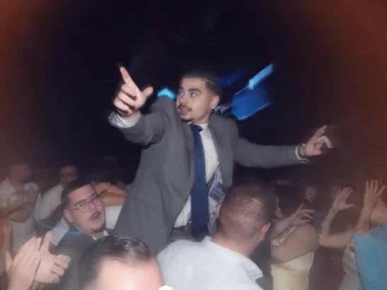 The Graduation Party: a young man in a suit lifted up on shoulders above the dancefloor, arm out, the crowd around him mid-cheer.